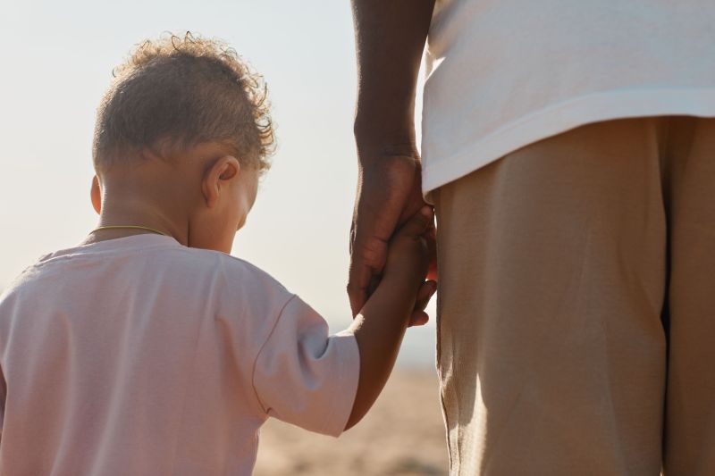 Back view portrait of cute African-American toddler holding hands with dad while enjoying walk on beach in sunlight, copy space